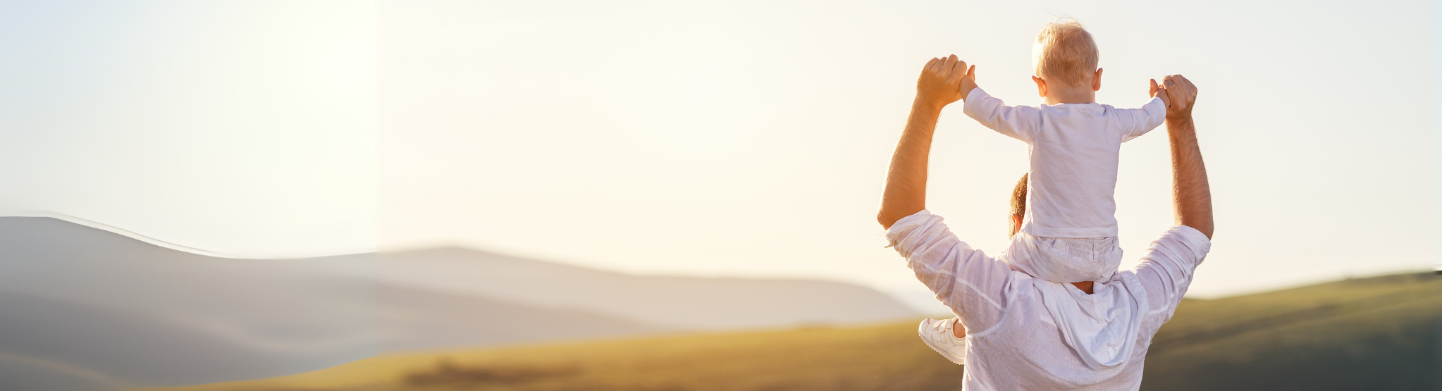 A father and son in white shirts standing on a hill at sunset.