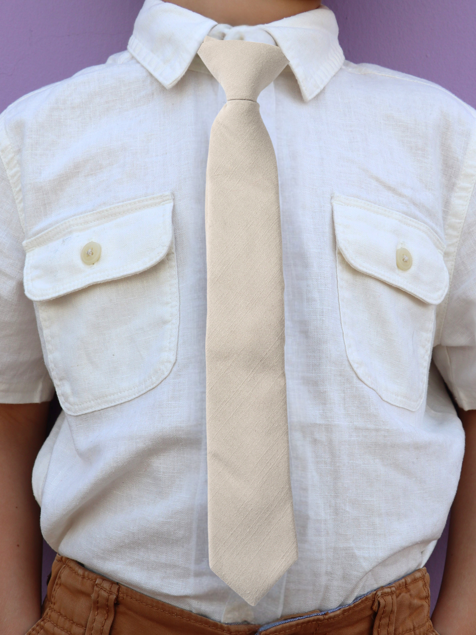 The torso of a boy wearing a solid kids tie with plain netural very light brown or sandy tan/gray  color.