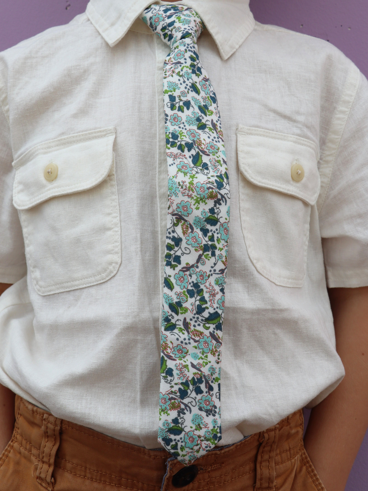 The torso of a boy wearing a floral kids tie with dusty blue flowers, pink accents, turquoise blossoms and bluish leaves.