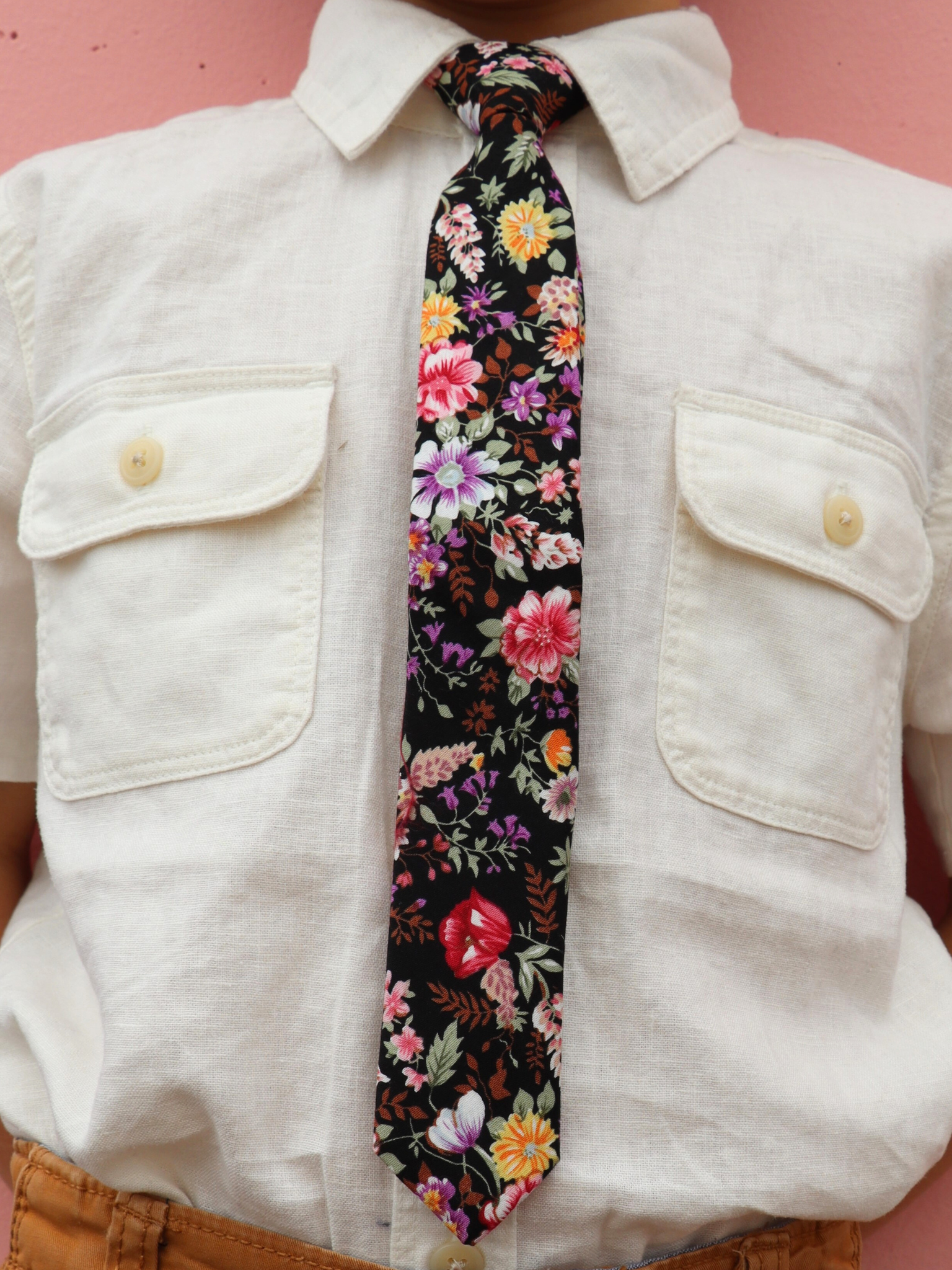 The torso of a boy wearing a floral kids tie with pink flowers, yellow petals, wisteria purple blossoms and brown leaves.