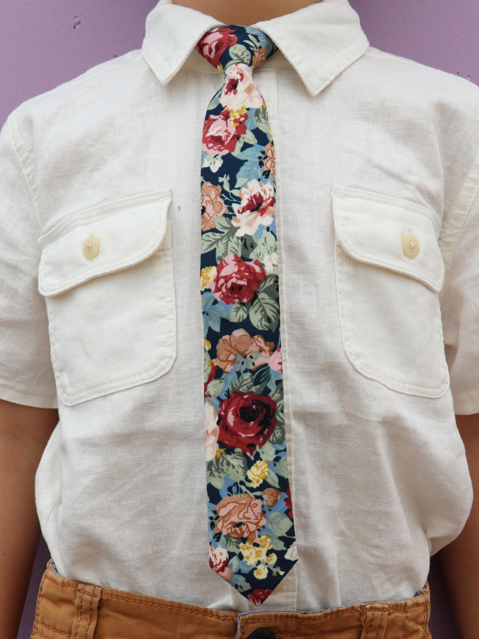 The torso of a boy wearing a floral kids tie with dark red flowers, burnt orange petals, roses and light green leaves.