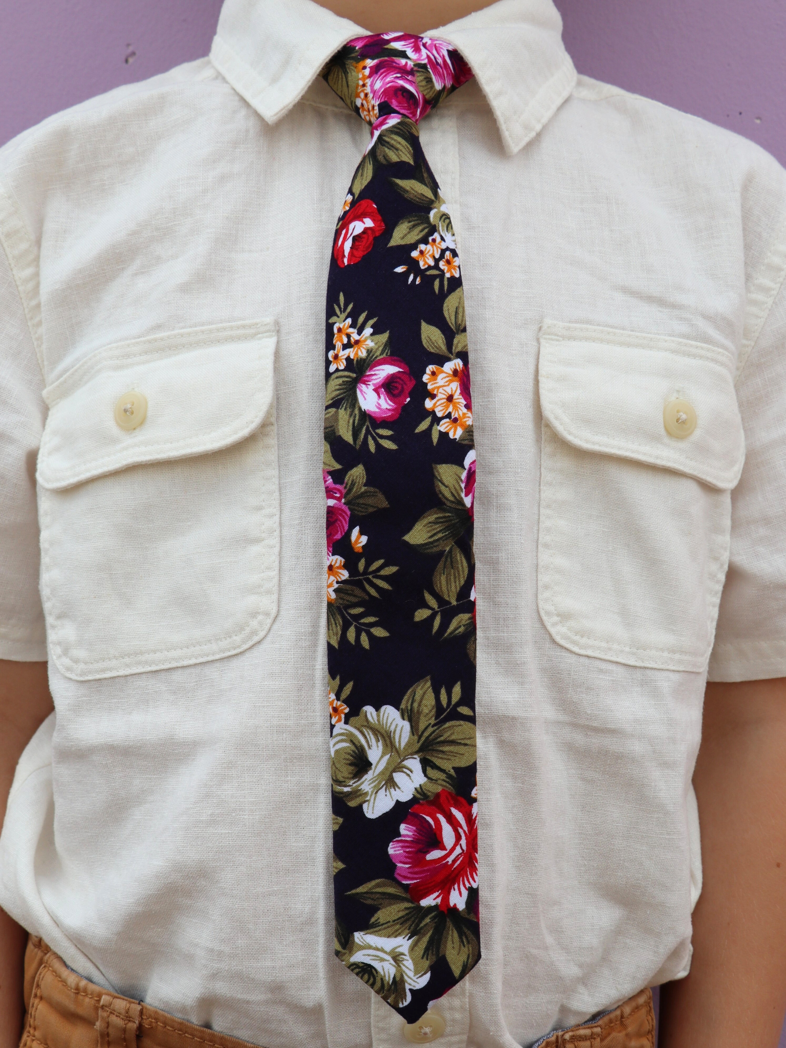 The torso of a boy wearing a floral kids tie with pink rose flowers, red petals, yellow blossoms and green leaves.