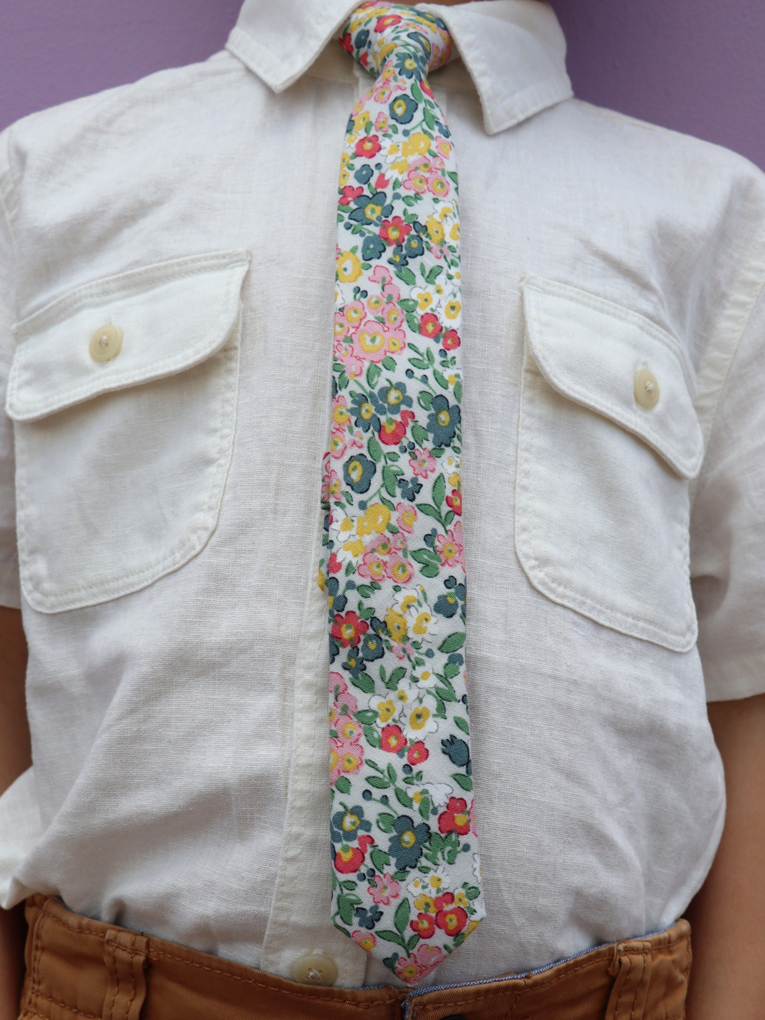 The torso of a boy wearing a floral kids tie with small pink flowers, teal petals, yellow blossoms and green leaves.