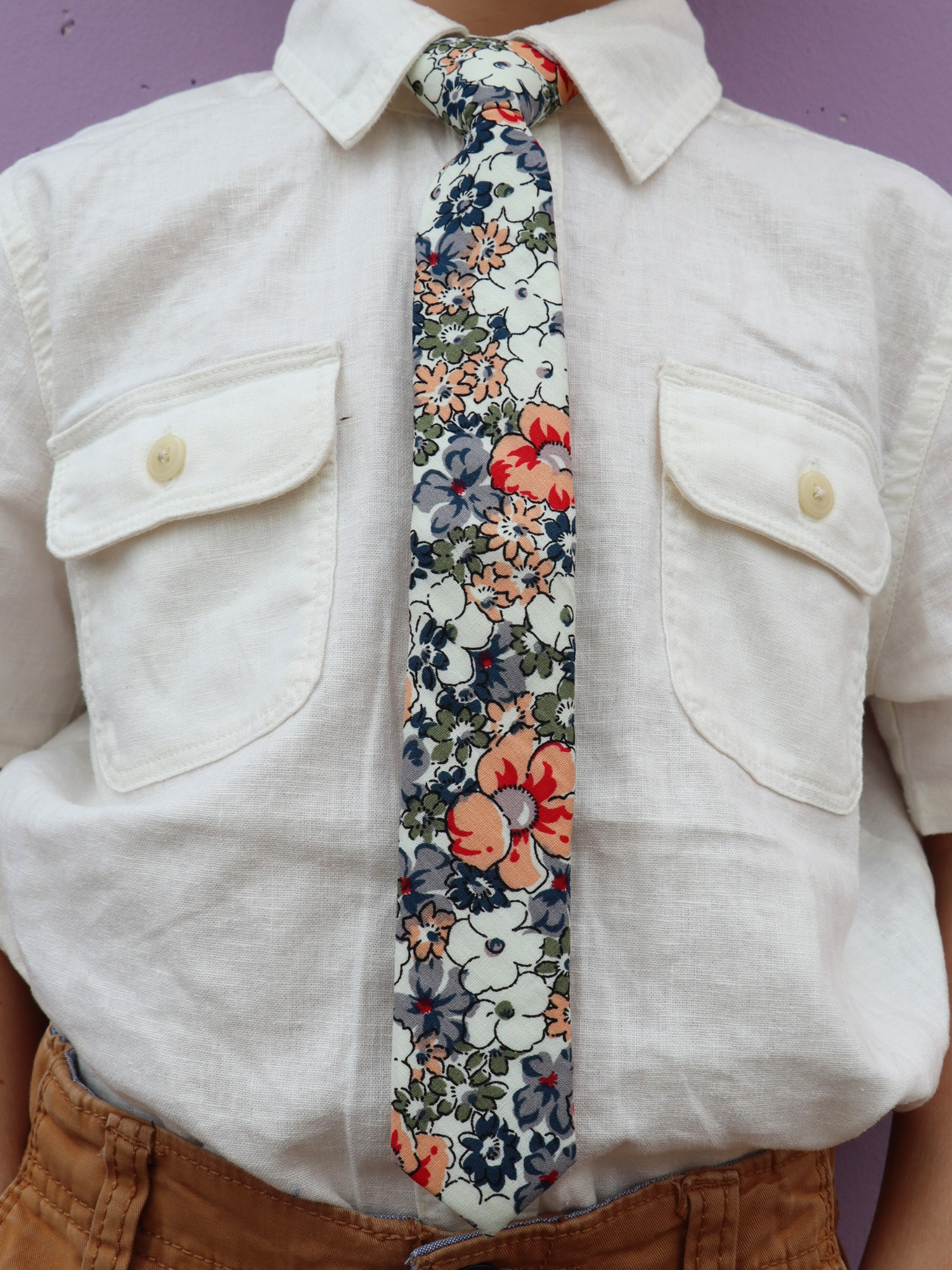 The torso of a boy wearing a floral kids tie with blue flowers, white petals, and peach or salmon blossoms and green leaves.