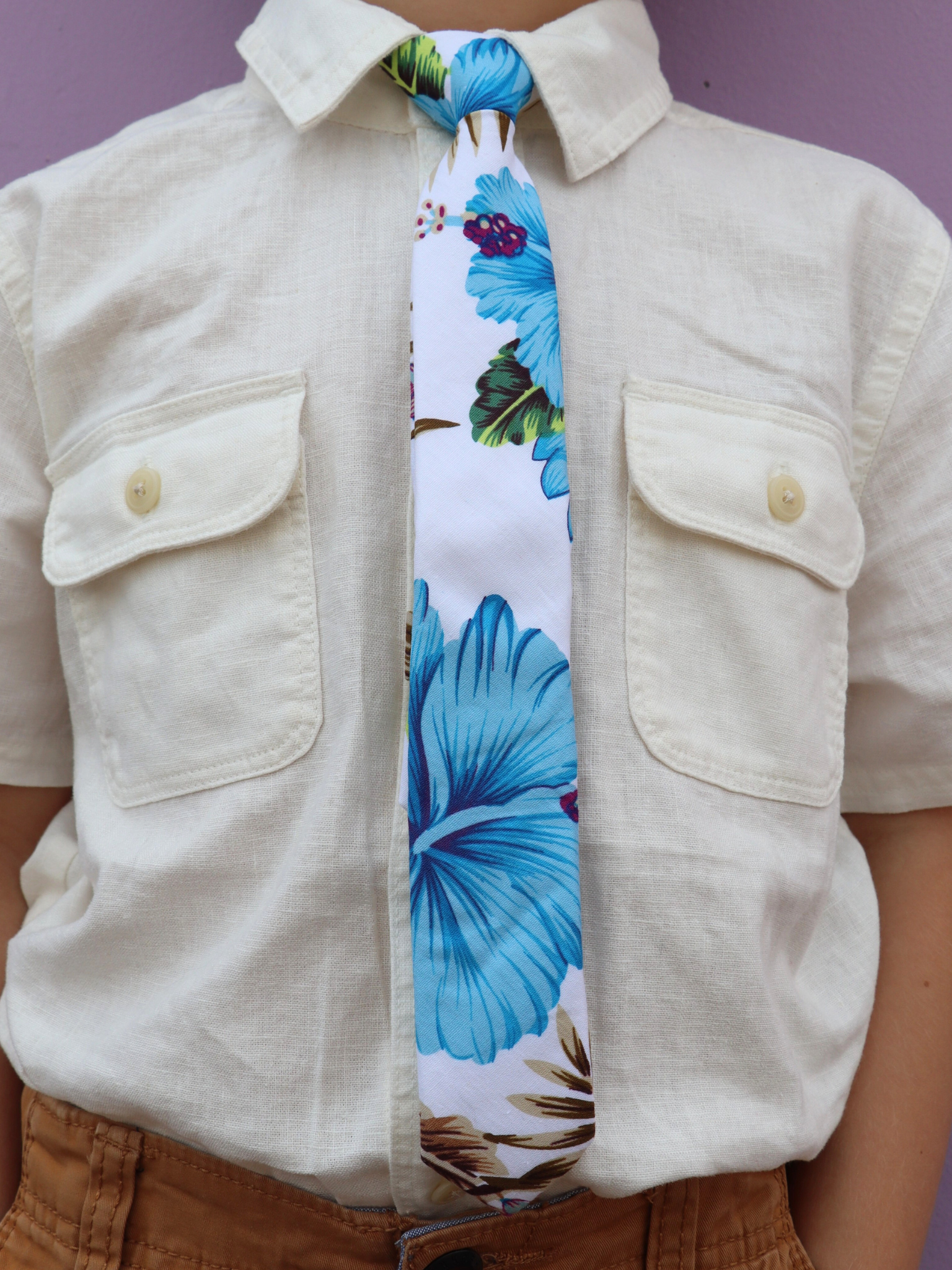 The torso of a boy wearing a floral kids tie with dusty blue beach flowers, hibiscus petals, spots of pink, and brown leaves.