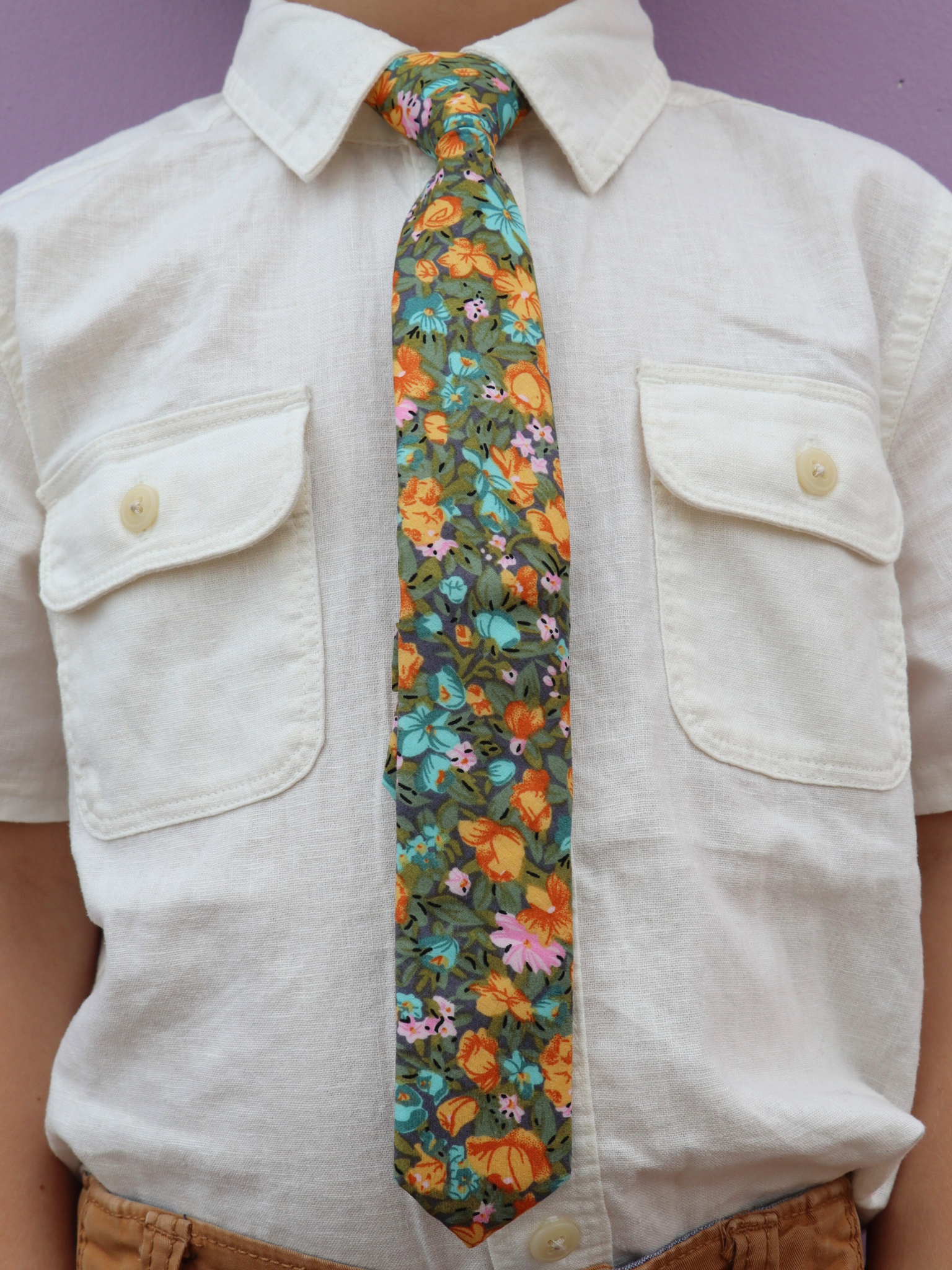 The torso of a boy wearing a floral kids tie with orange flowers, dark pletals, light blue blossoms and green leaves.