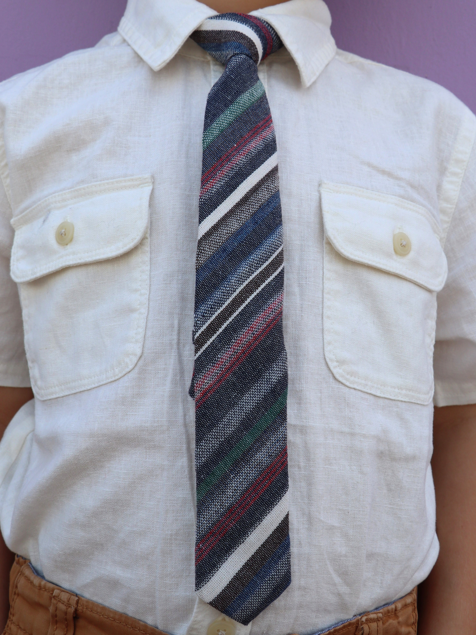 The torso of a boy wearing a striped kids tie with dark gray stripes, thin red lines, light grey bars and diagonal designs.