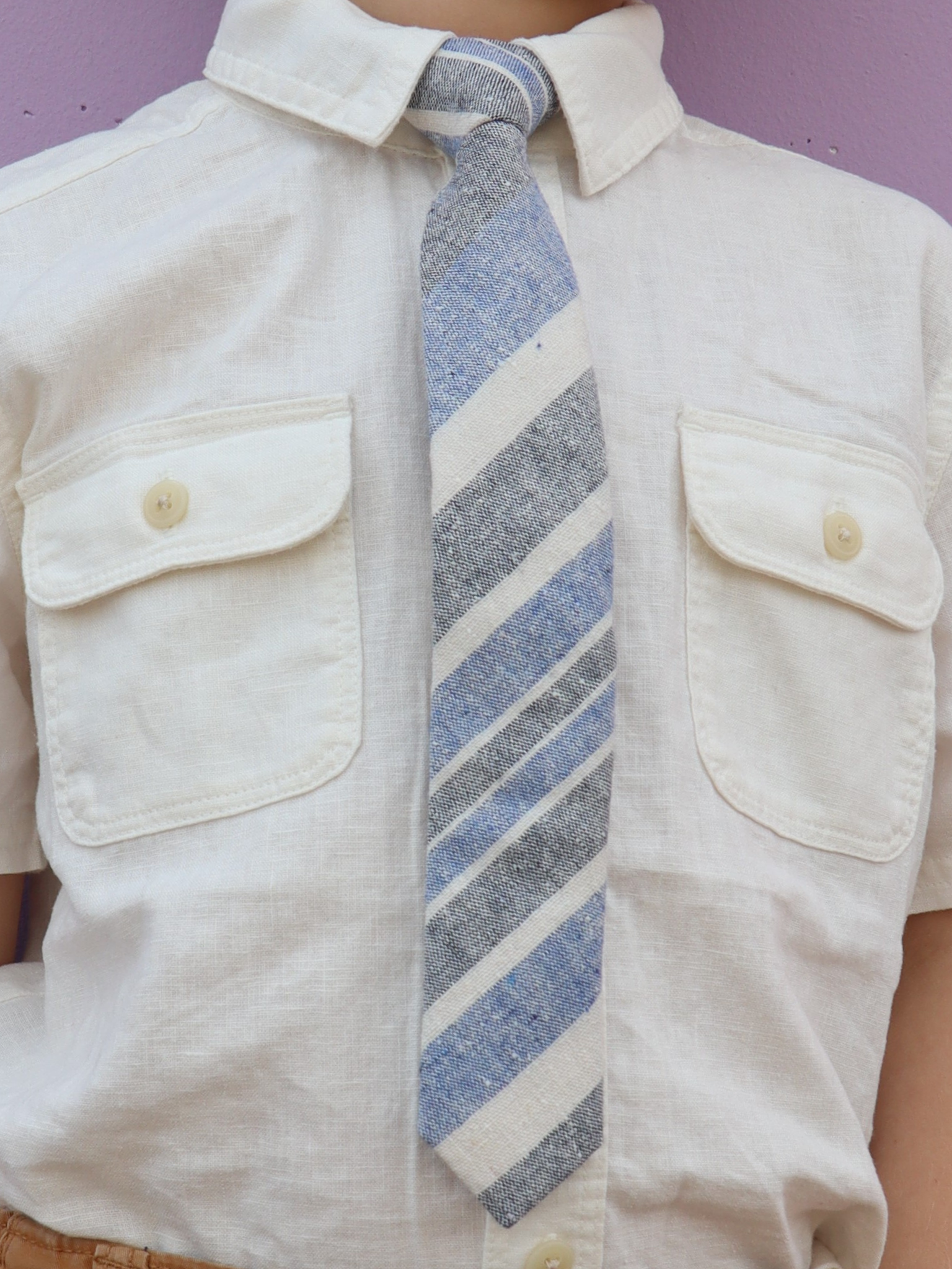 The torso of a boy wearing a striped kids tie with denim blue stripes, beige cream lines, grey bars and diagonal designs.