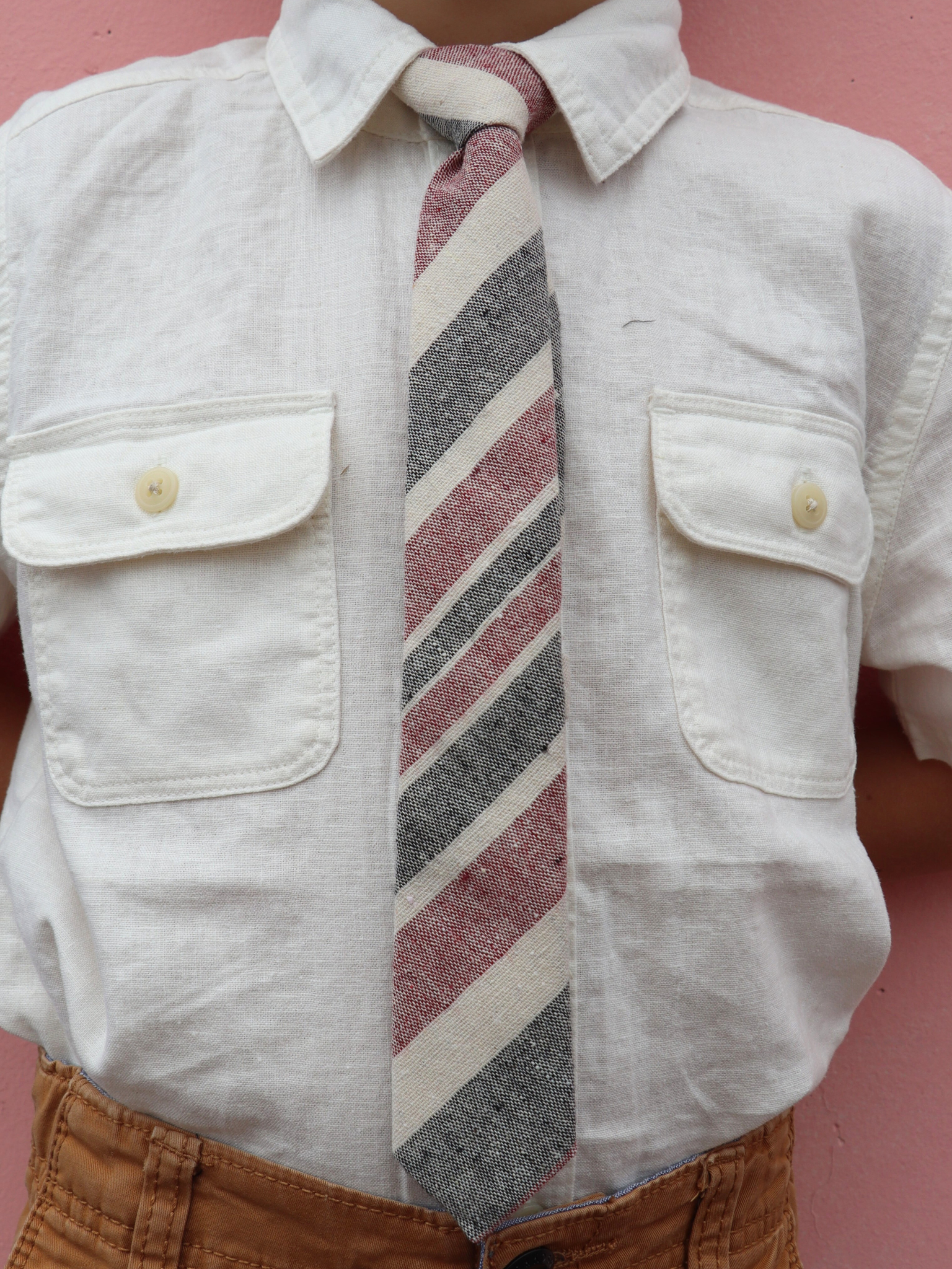 The torso of a boy wearing a striped kids tie with crimson red stripes, dark gray lines, beige bars and diagonal designs.