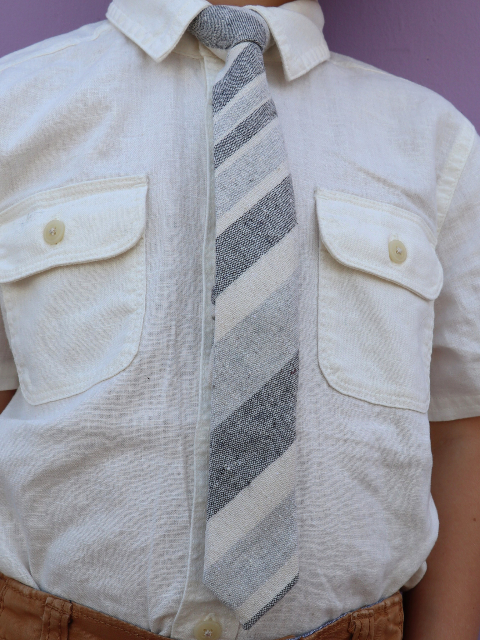 The torso of a boy wearing a striped kids tie with pale grey stripes, dark gray lines, cream  bars and diagonal designs.