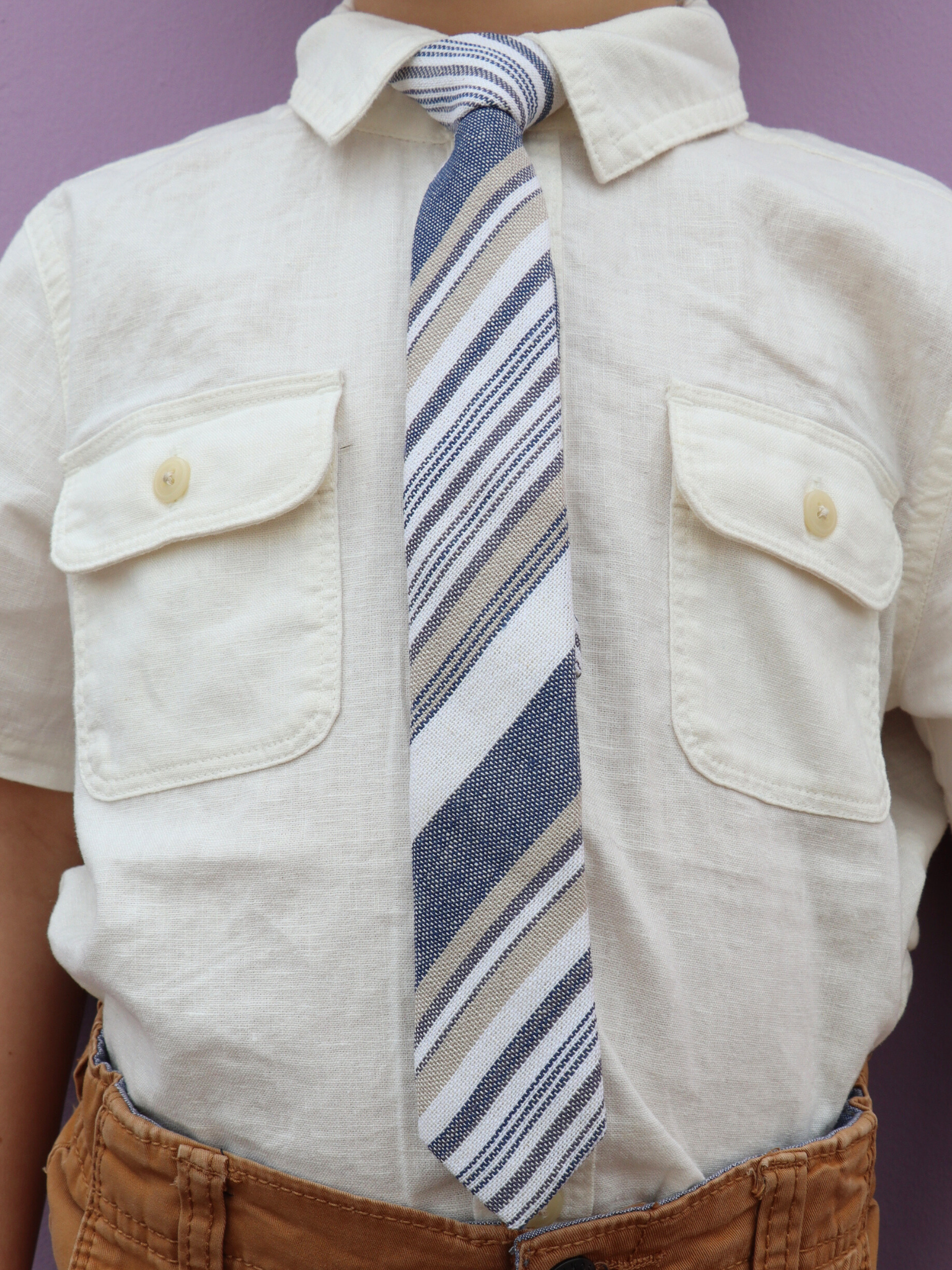 The torso of a boy wearing a striped kids tie with thin dark blue stripes, white lines, gold bars and diagonal designs.