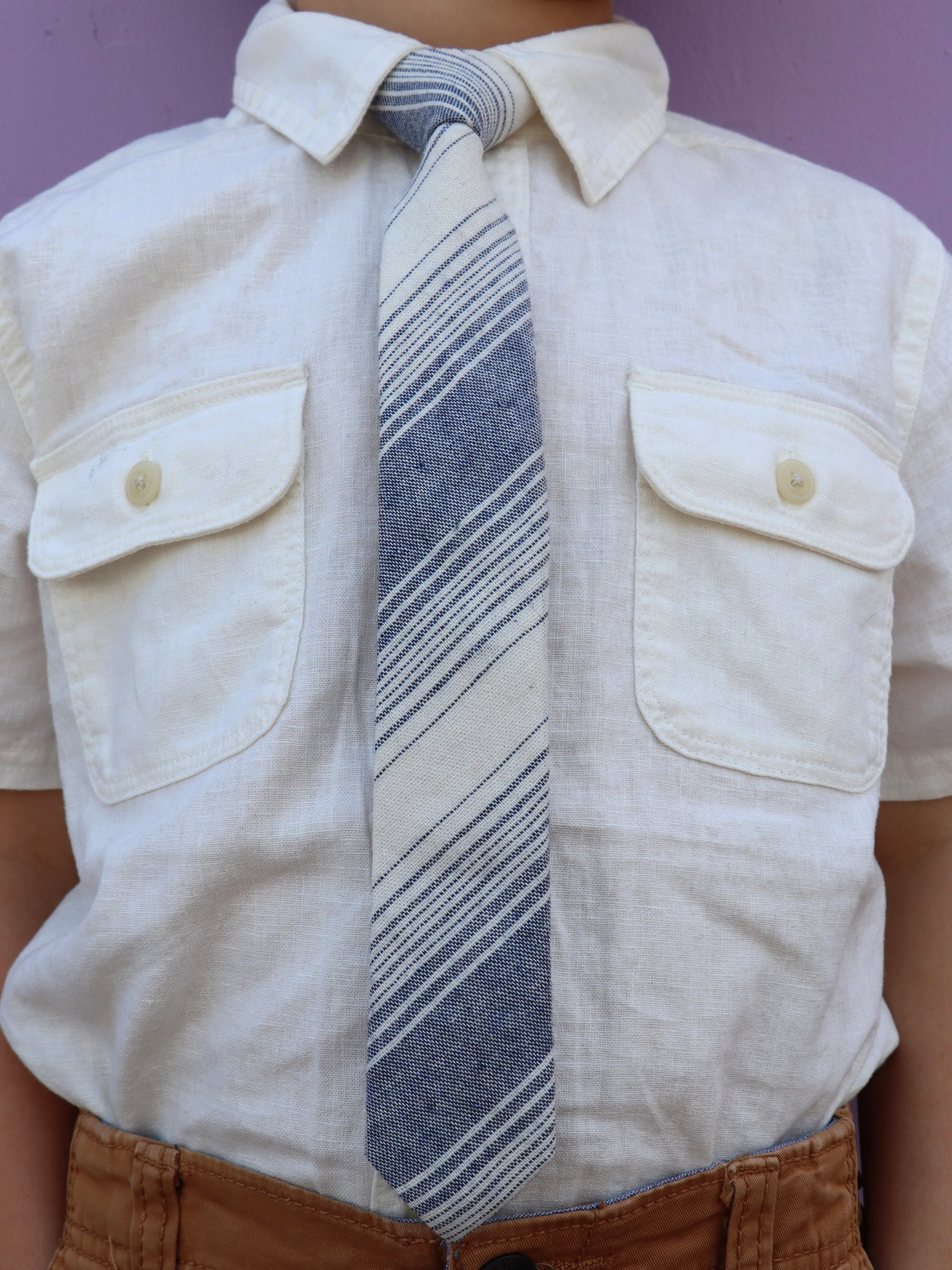 The torso of a boy wearing a striped kids tie with navy blue stripes, thin beige cream lines,  and diagonal designs.