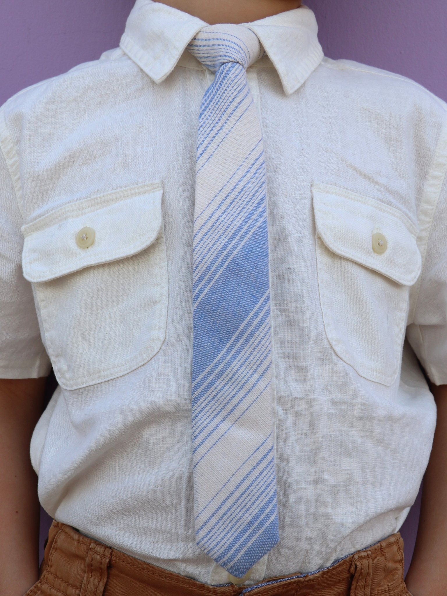 The torso of a boy wearing a striped kids tie with light dusty blue stripes, thin beige cream lines, and off-white.