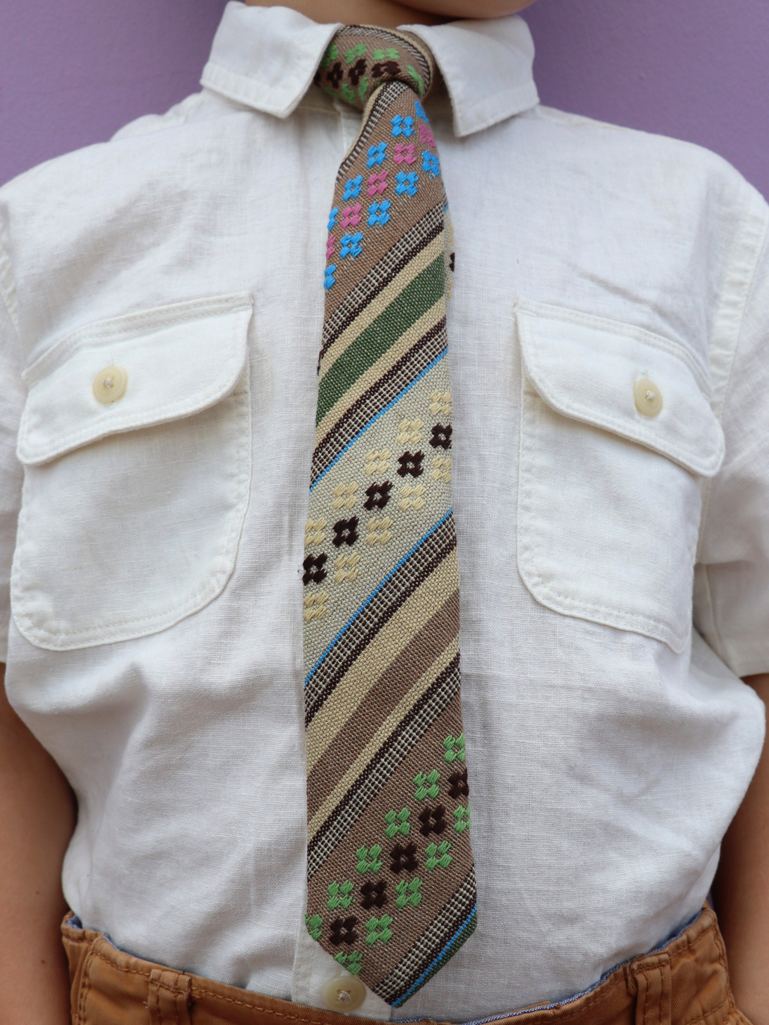 The torso of a boy wearing a striped kids tie with green stripes, tan lines, brown bars and cross designs.