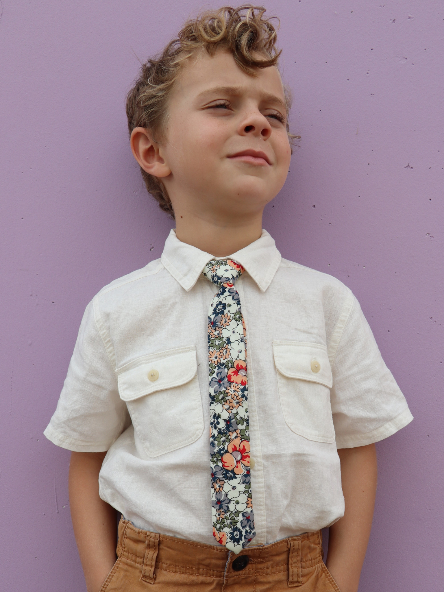 A boy in a white shirt with a peach, white, and blue floral pre tied tie for kids ages 1-12 available in 4 childrens sizes.