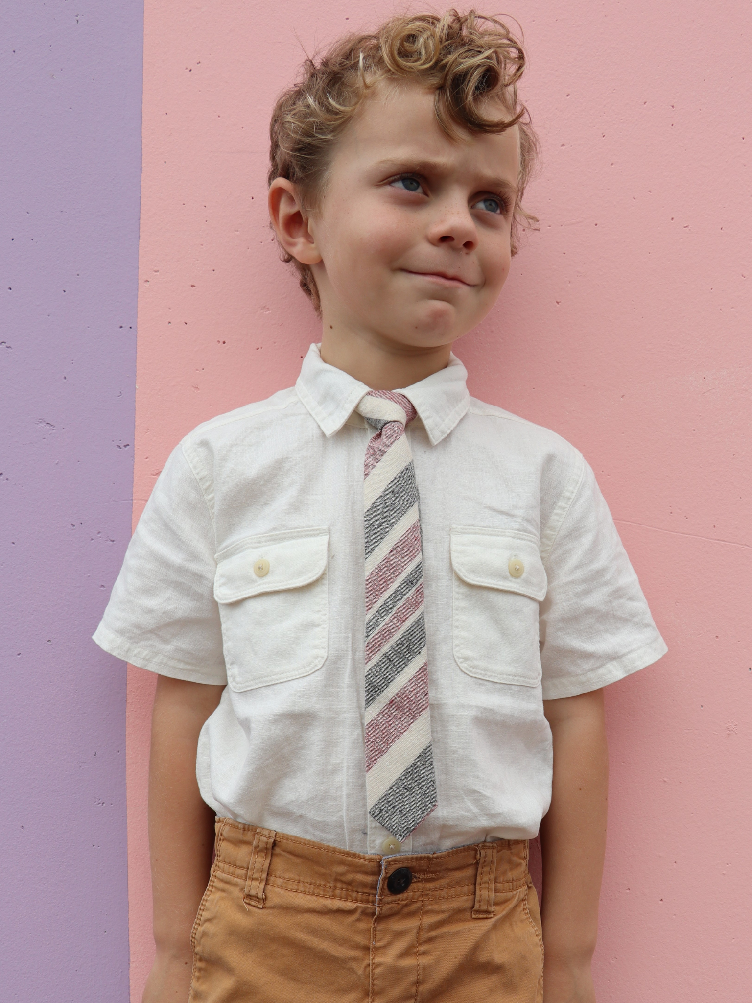 A boy in a white shirt with a red, gray, and beige striped pre tied tie for kids ages 1-12 available in 4 childrens sizes.