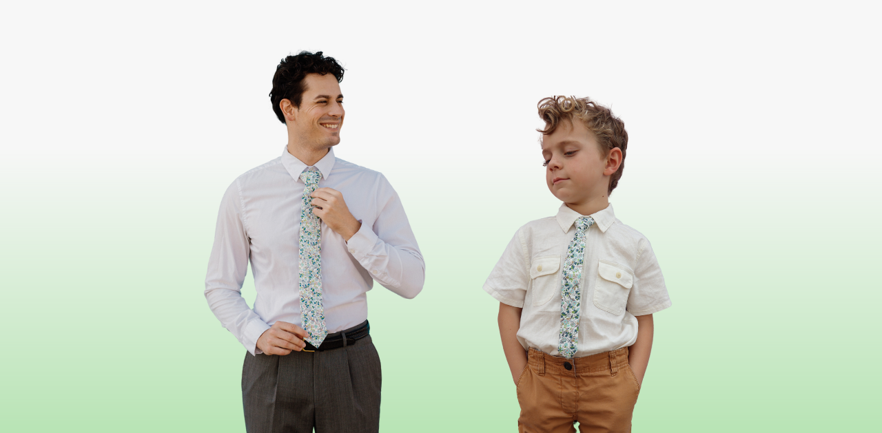 A man and boy in matching blue and green floral ties by Otis and Oliver standing in front of a green background. 